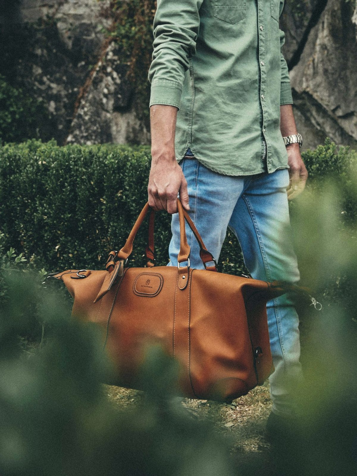 A man carrying a cognac leather weekender through a garden